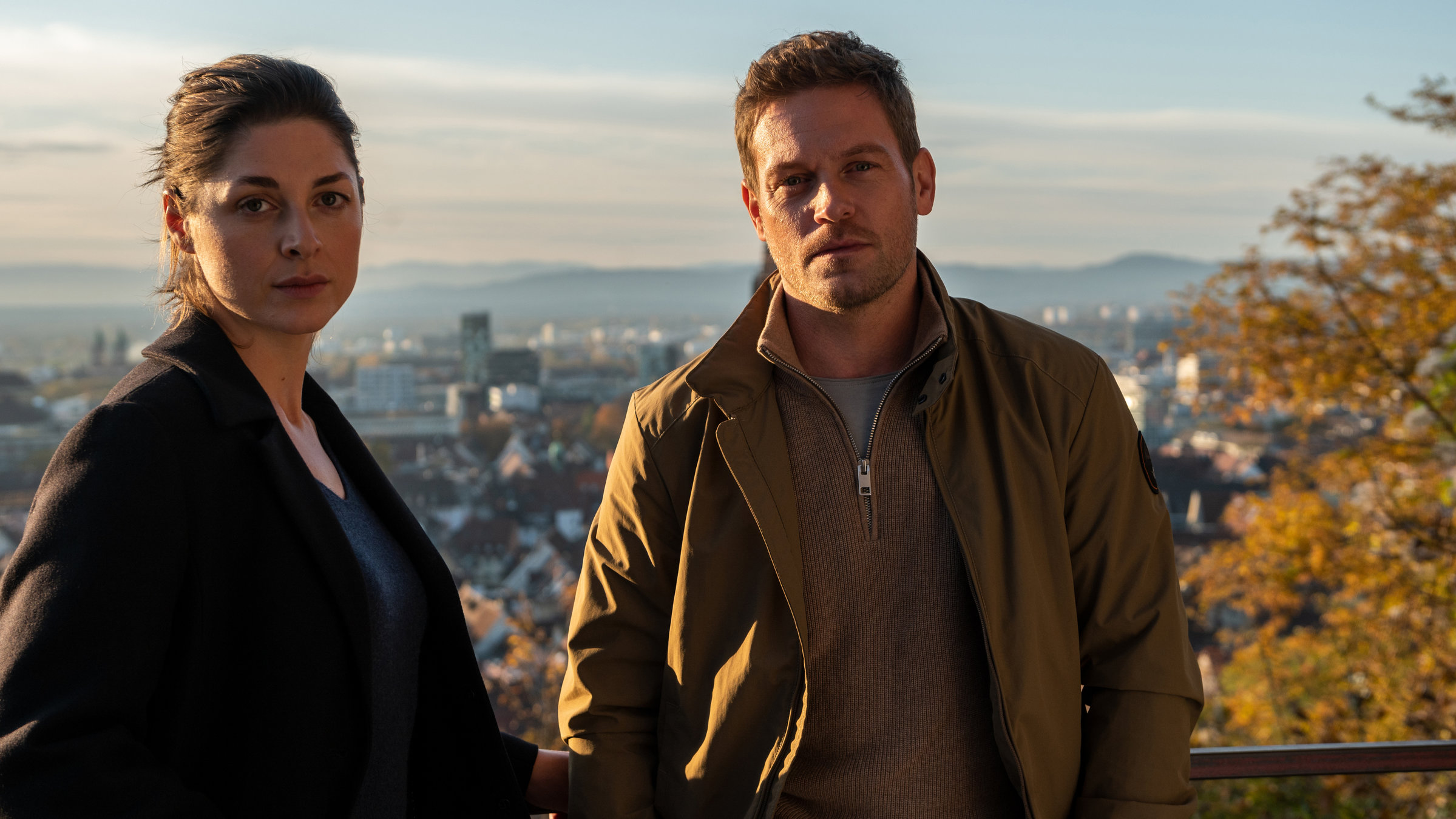 "Breisgau - Nehmen und Geben": Tanja Wilken (Katharina Nesytowa) und Dennis Danzeisen (Joscha Kiefer) stehen am Geländer eines Aussichtspunkts und blicken entschlossen in die Kamera. Hinter ihnen ist das Stadtpanorama von Freiburg zu sehen.