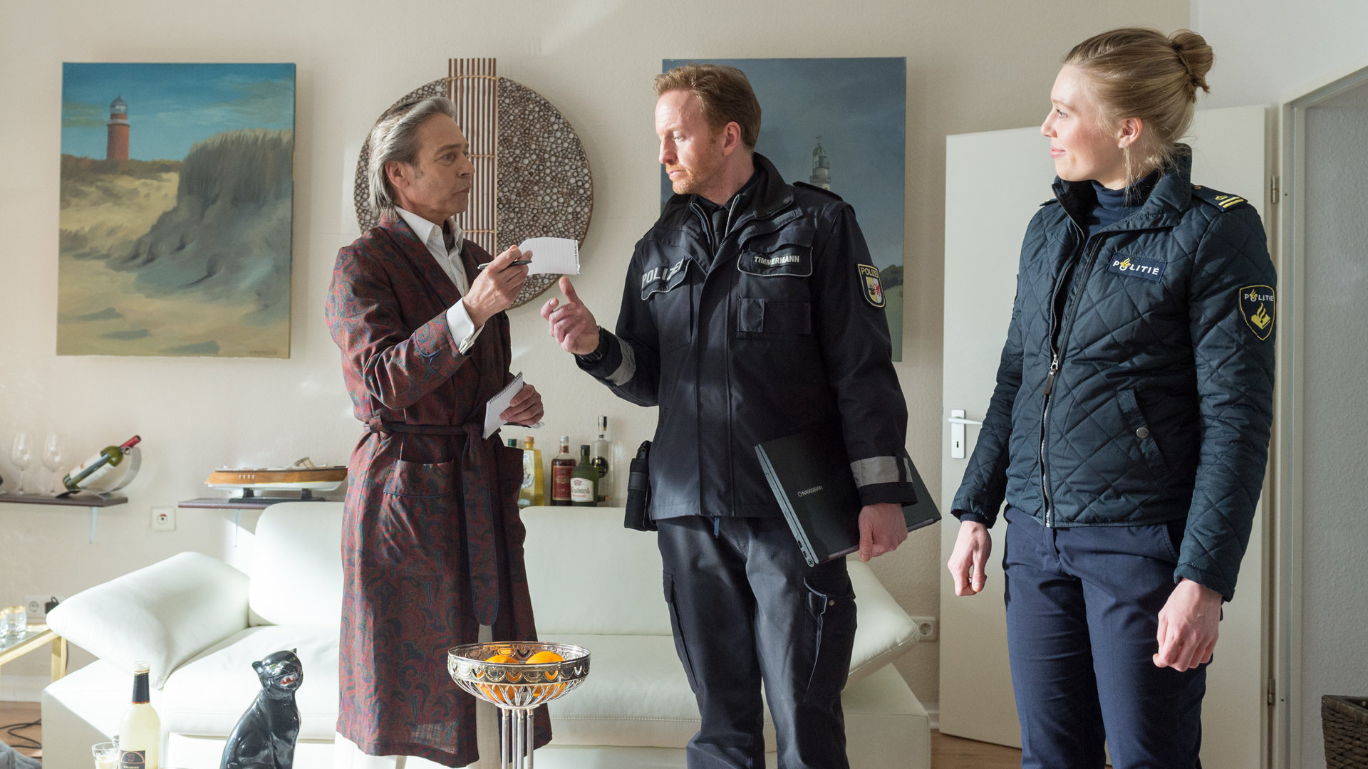 "SOKO Wismar – Der Eintänzer": Hubertus Kalow (Jaques Breuer) steht im Morgenmantel in seinem Wohnzimmer. Neben ihm stehen Kai Timmermann (Mathias Junge) und Anneke van der Meer (Isabel Berghout) in Uniform und sehen zu ihm herüber. Timmermann hat einen Laptop unter dem Arm und nimmt einen kleinen Zettel entgegen, den Kalow ihm reicht.