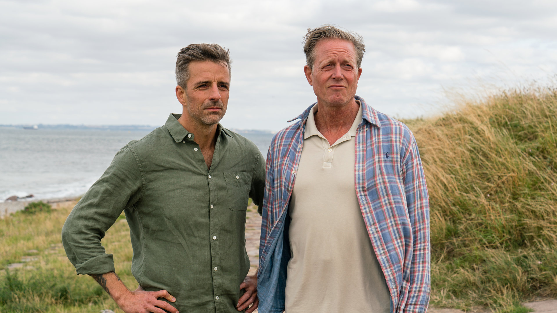 "Dan Sommerdahl - Tödliche Idylle: Liebeslabyrinth": Flemming Torp (André Babikian) und Dan Sommerdahl (Peter Mygind) stehen am Meer vor einer Düne. Es ist windig.