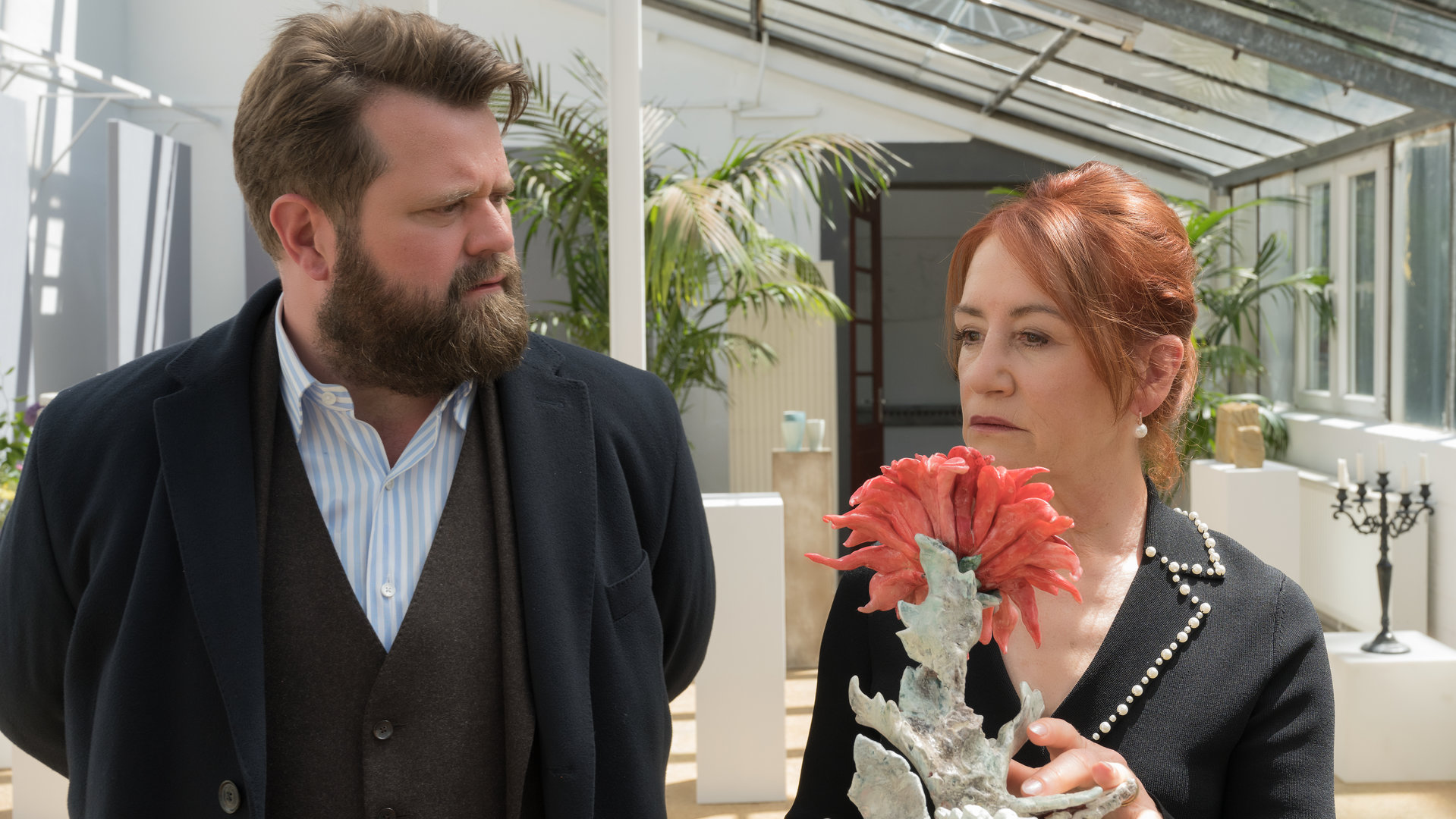 "Ein Fall für zwei - In Liebe W.": Benni Hornberg (Antoine Monot, Jr.) steht mit Marianne Geiger (Ulrike Krumbiegel) im Ausstellungsraum, der sich in einem Wintergarten befindet. Vor ihnen steht eine Skulptur einer Dahlie. Die beiden unterhalten sich.