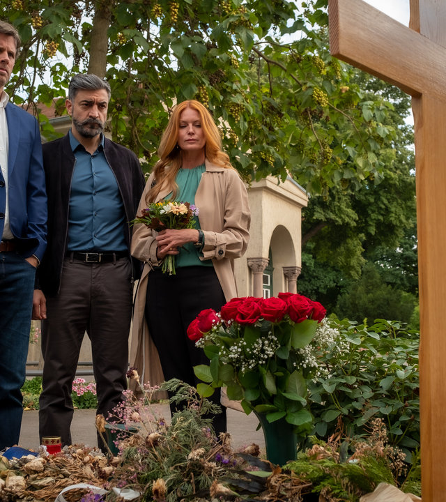 "Blutige Anfänger - Frauenmörder": Pierre Hartwich (Steffen Groth), Sami Malouf (Neil Malik Abdullah) und Julia Salomon (Esther Schweins) stehen nebeneinander vor einem Grab. Julia hält einen bunten Blumenstrauß in den Händen. Auf dem Grab steht ein frischer Strauß rote Rosen.