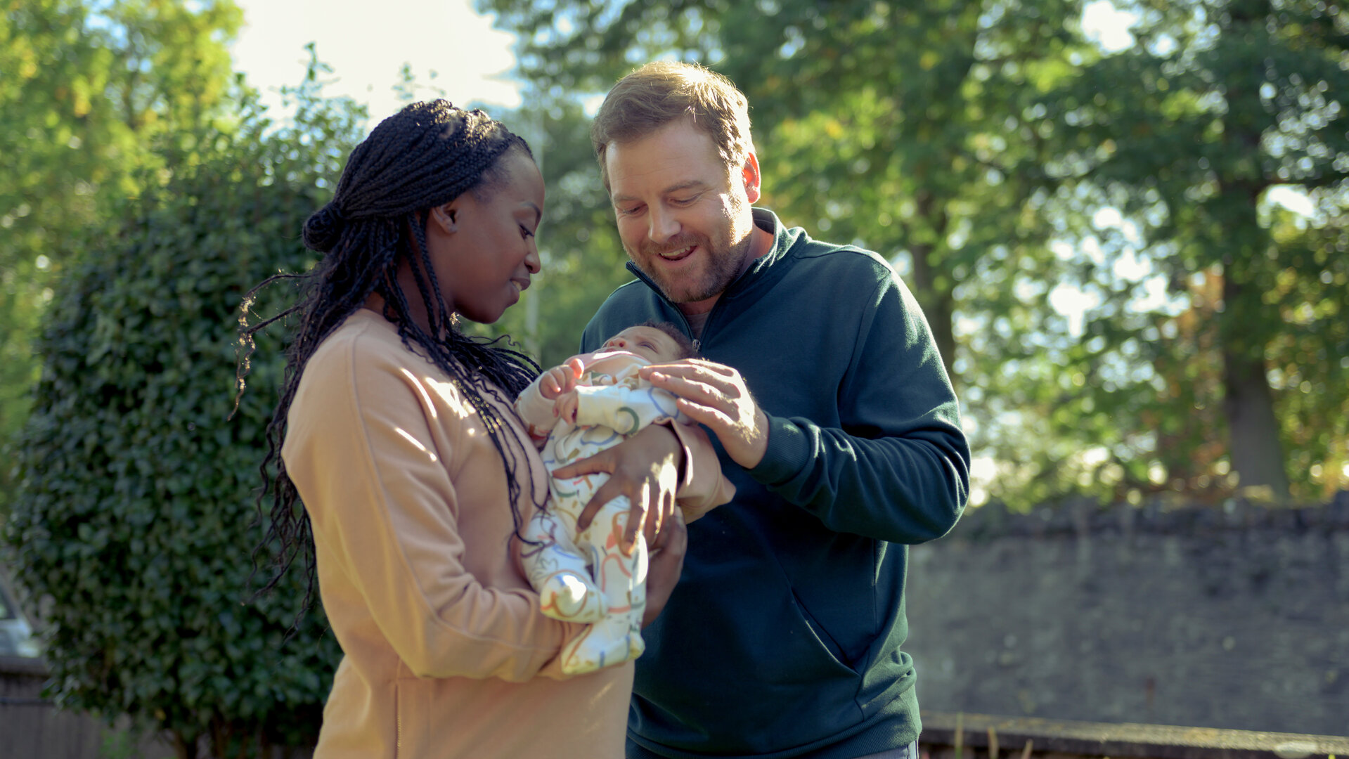 "Northern Lights - Polarlichter": Lloyd (Stephen Jones) und Denise (Jennifer Heylen) stehen nebeneinander im Freien. Sie hält ein kleines Kind im Strampelanzug im Arm. Er fasst es behutsam an.