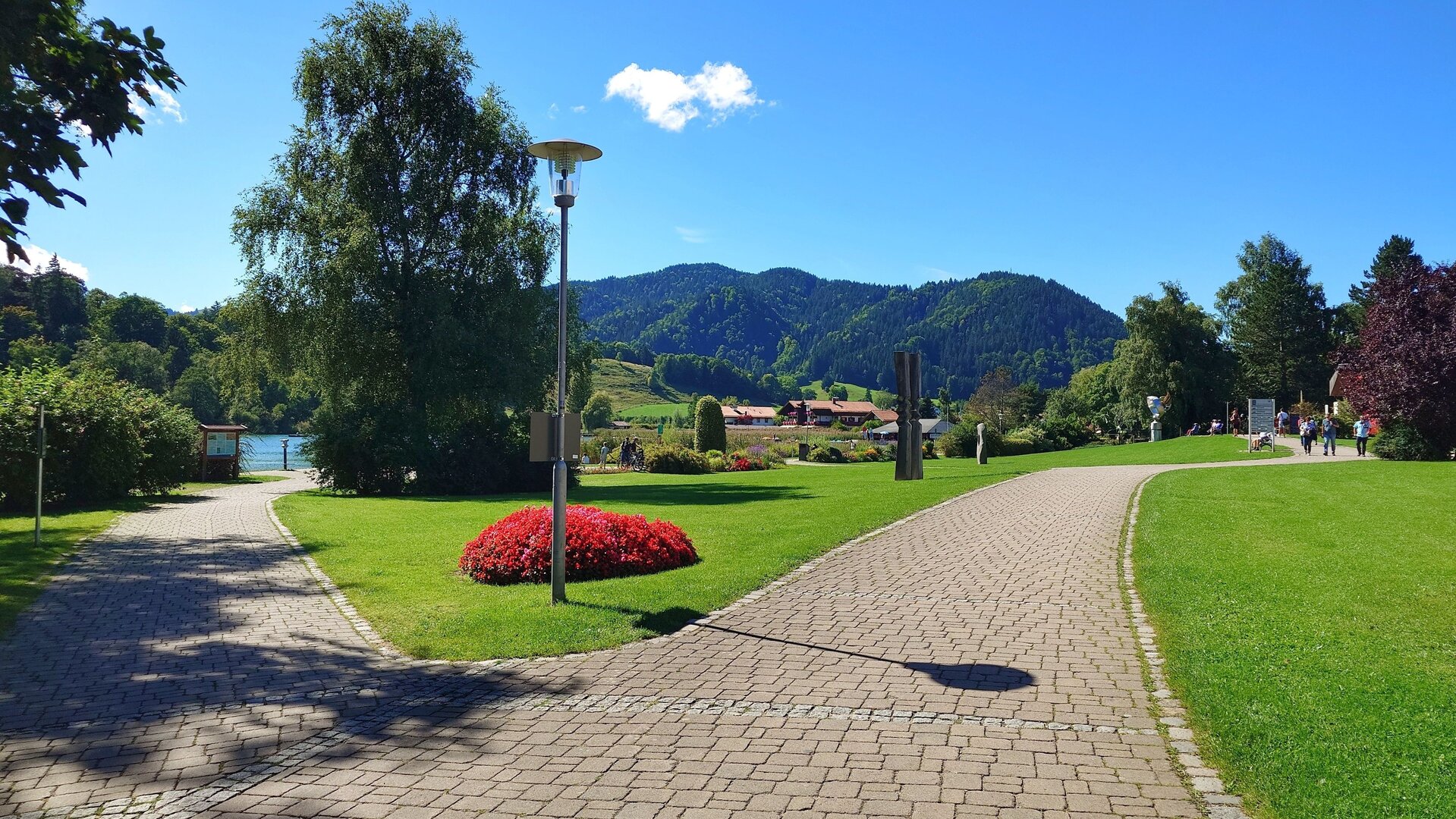 "Evangelischer Open-Air-Gottesdienst: Kopfüber ins Leben": Kurpark am Schliersee: In einer gepflegten Grünanlage gabeln sich zwei Wege im Vordergrund. Ein Weg führt zum Schliersee. Im Hintergrund ragen grüne Berge in den blauen Himmel.