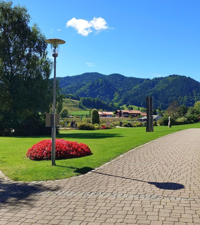 "Evangelischer Open-Air-Gottesdienst: Kopfüber ins Leben": Kurpark am Schliersee: In einer gepflegten Grünanlage gabeln sich zwei Wege im Vordergrund. Ein Weg führt zum Schliersee. Im Hintergrund ragen grüne Berge in den blauen Himmel.