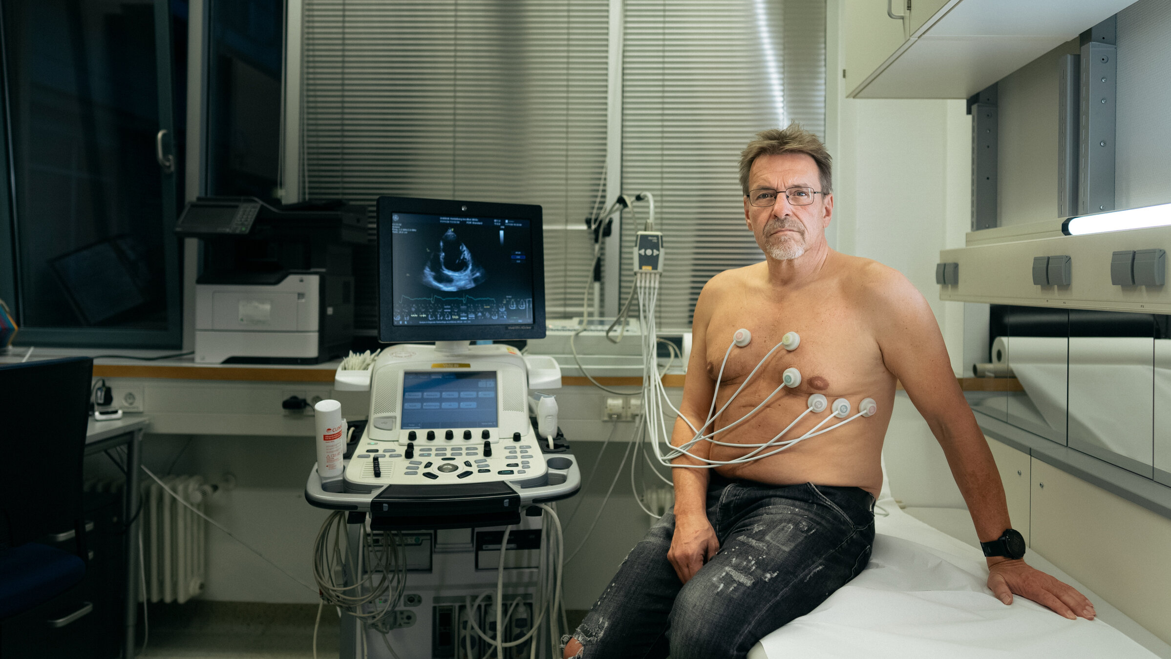 "Mein widerspenstiges Herz": Harald sitzt beim Arzt. Elektroden kleben auf seinem nackten Oberkörper. Auf dem Monitor neben ihm sieht man ein Ultraschallbild seines Herzens.