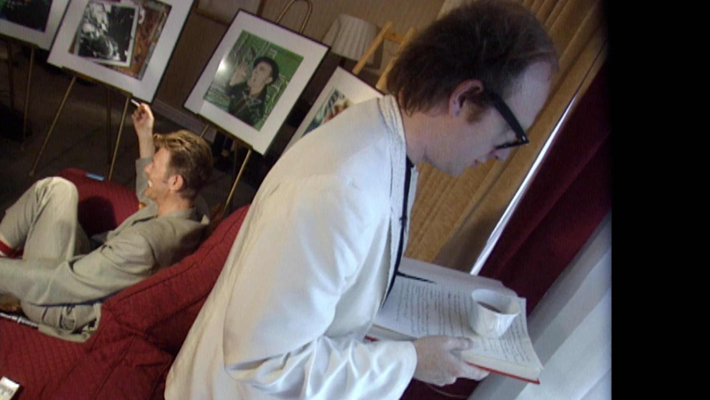 "Why are we creative": Mann auf dem Sofa sitzend guckt nach links, anderer Mann steht rechts im Bild mit Buch in der Hand mit dem Rücken zum Gesprächspartner.