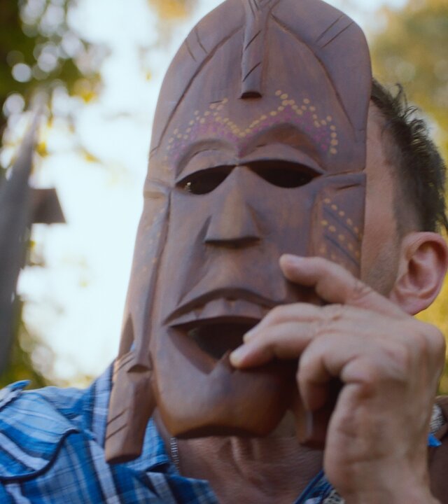 "Doppelhaushälfte - Cultural Heritage": Andi (Milan Peschel) hält eine geschnitzte Maske aus Holz vor sein Gesicht. Die Maske hat schmale Augenöffnungen und einen weit geöffneten Mund. Im Vordergrund ist eine Speerspitze zu sehen.