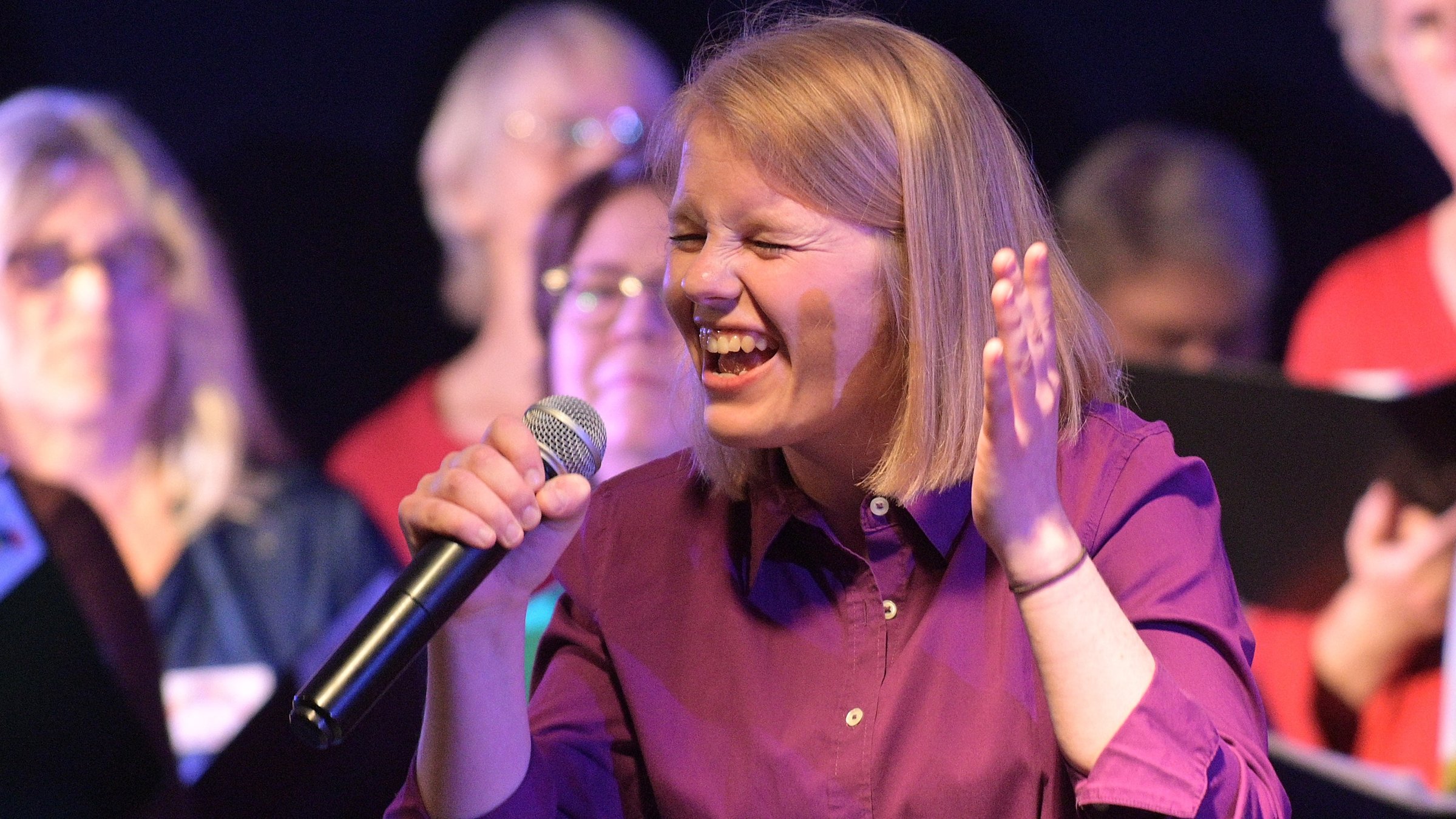 "37°: Singen fürs Leben – Im Chor gegen den Krebs": Nahaufnahme. Julja hält in der rechten Hand ein Mikrofon in Höhe des Mundes und singt mit geschlossenen Augen. Sie hat schulterlanges blondes Haar und trägt eine lilafarbene Bluse. Im Hintergrund stehen Sängerinnen und Sänger des Chors.