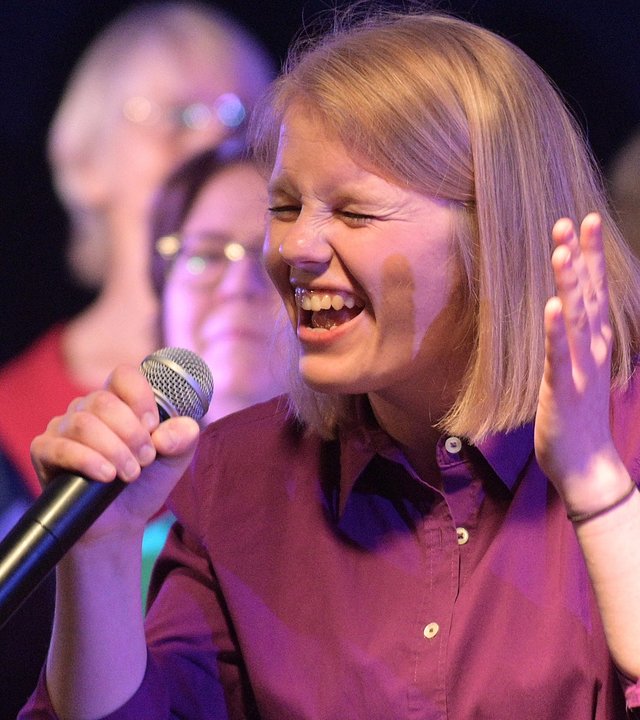 "37°: Singen fürs Leben – Im Chor gegen den Krebs": Nahaufnahme. Julja hält in der rechten Hand ein Mikrofon in Höhe des Mundes und singt mit geschlossenen Augen. Sie hat schulterlanges blondes Haar und trägt eine lilafarbene Bluse. Im Hintergrund stehen Sängerinnen und Sänger des Chors.