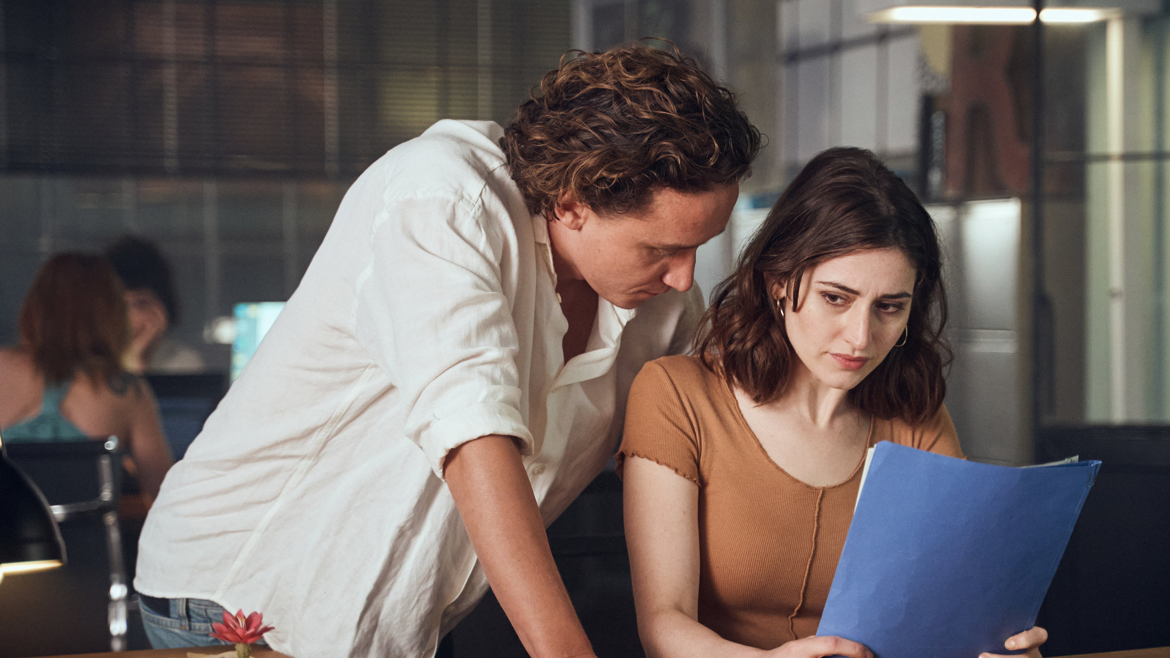"Der Schatten - Wunderland": Emil Fermus (Aaron Friesz) lehnt auf dem Tisch und schaut Norah Richter (Deleila Piasko) über die Schulter. Diese hält eine blaue Mappe in ihrer Hand. Vor ihr auf dem Tisch stehen Schreibartikel und ein Kaktus. Im Hintergrund sieht man einen Büroraum.