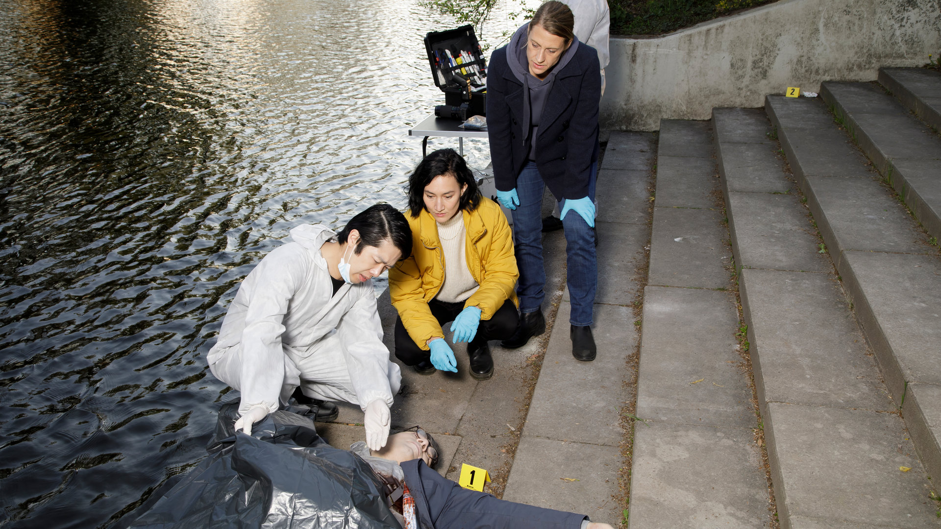 "SOKO Potsdam - Unter Kontrolle": Tamara Meurer (Anja Pahl) und Pauline Hobrecht (Agnes Decker) sind am Tatort beim Fluss an einer Treppe. Thomas Brandner (Yung Ngo) hockt neben ihnen vor der Leiche von Vadim Pasetzki (Komparse), an dessen Brust viel Blut zu sehen ist.