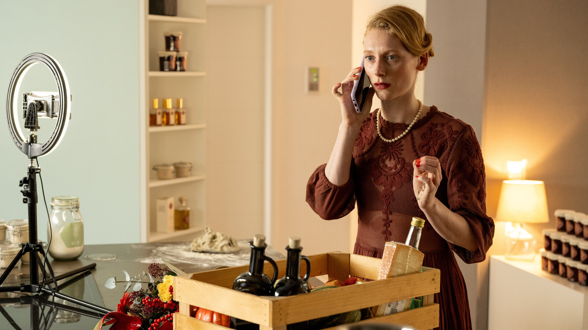"SOKO Köln - Tradwife": Sofia Jansen (Dana Herfurth) steht an einem Tisch und hält sich mit ernstem Blick ihr Handy ans Ohr. Vor ihr steht eine braune Kiste, in der sich Lebensmittel befinden. Neben der Kiste steht ein Ringlicht, in dem eine Kamera befestigt ist. Im Hintergrund stehen in einem geöffneten Schrank Kosmetikartikel.
