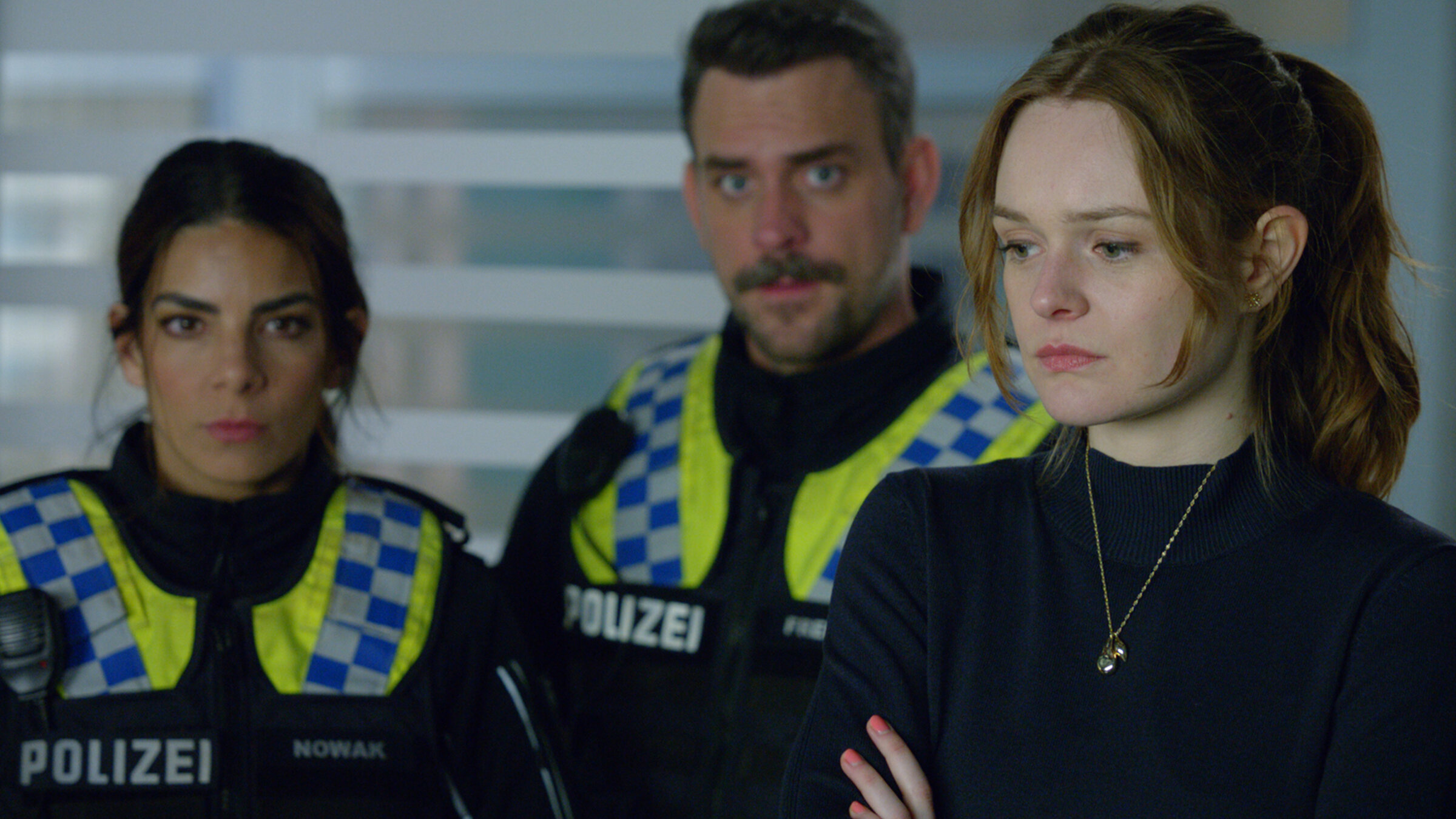 "Notruf Hafenkante - Schweres Erbe": Großaufnahme von Johanna Severin (Miriam Schiweck). Sie steht im Krankenzimmer ihres Vaters und schaut nachdenklich. Seitlich hinter ihr stehen die Polizistin Isa (Lilli Hollunder) und ihr Kollege Kris (Marc Barthel). Beide blicken ebenso nachdenklich.