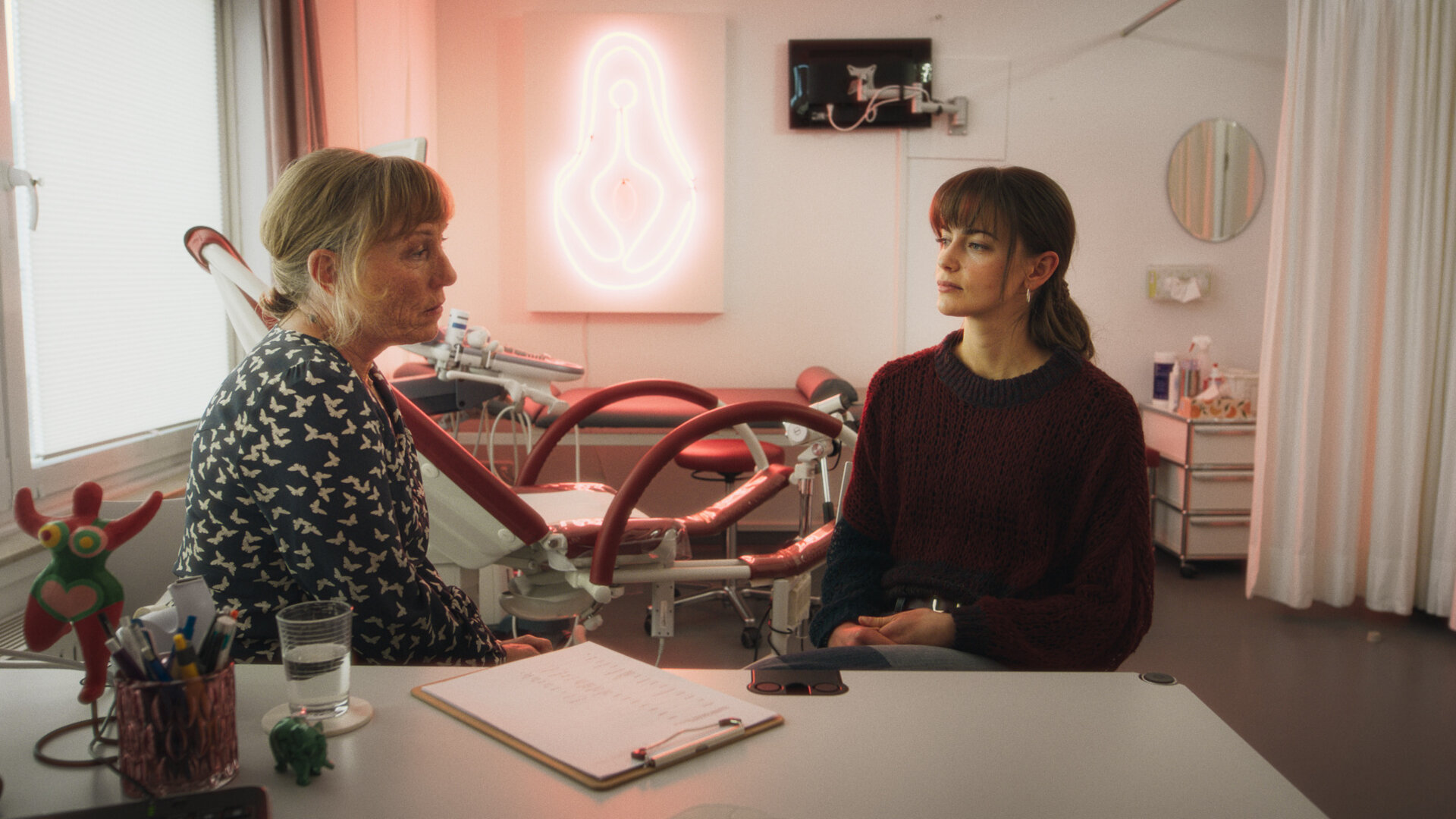 "Bauchgefühl - Dr. Roithner": Dr. Roithner (Antje Lewald) und Lena (Laura Berlin) sitzen sich in Dr. Roithners Praxis auf der selben Seite des Schreibtisches gegenüber. Lena knetet ihre Finger im Schoß. Die Frauen schauen sich an. Auf dem Tisch liegt ein Klemmbrett mit Fragebogen. Daneben steht ein Glas Wasser, eines mit Stiften und eine bunte Frauenskulptur.