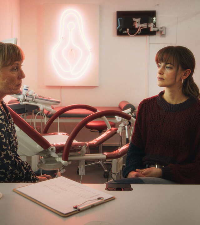 "Bauchgefühl - Dr. Roithner": Dr. Roithner (Antje Lewald) und Lena (Laura Berlin) sitzen sich in Dr. Roithners Praxis auf der selben Seite des Schreibtisches gegenüber. Lena knetet ihre Finger im Schoß. Die Frauen schauen sich an. Auf dem Tisch liegt ein Klemmbrett mit Fragebogen. Daneben steht ein Glas Wasser, eines mit Stiften und eine bunte Frauenskulptur.