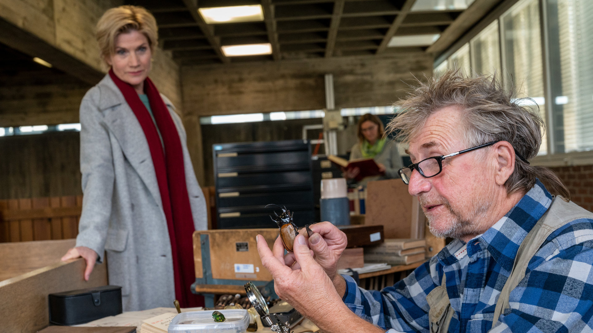 "SOKO Stuttgart – Letztes Kapitel": Klaus Michael Stoirer (Martin Semmelrogge) sitzt in der Bücherei an einem Schreibtisch, während er in der einen Hand einen Käfer und in der anderen eine Pinzette hält. Er schaut den Käfer fasziniert und konzentriert an, während sich Hauptkommissarin Martina Seiffert (Astrid M. Fünderich) an das Büchereigeländer lehnt und den Hobby-Käferspezialisten bei seiner Arbeit beobachtet.
