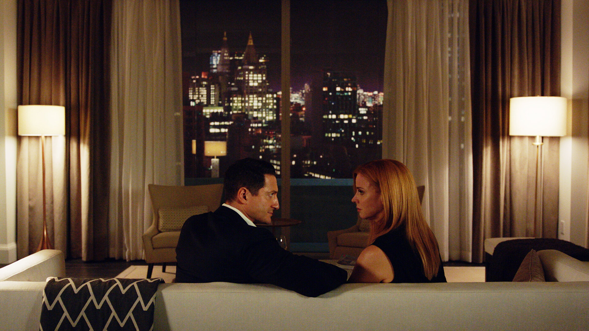 "Suits - Der Lockvogel": Thomas (Sasha Roiz) und Donna (Sarah Rafferty) sitzen auf einem cremefarbenen Sofa und schauen sich intensiv an. Im Bildhintergrund ist durch ein Fenster mit cremefarbenen Vorhängen die Skyline New Yorks zu sehen.