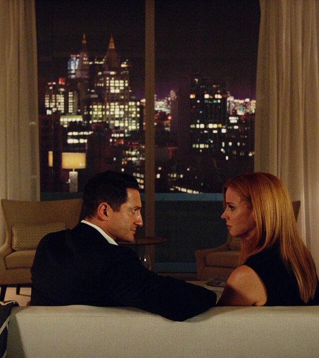 "Suits - Der Lockvogel": Thomas (Sasha Roiz) und Donna (Sarah Rafferty) sitzen auf einem cremefarbenen Sofa und schauen sich intensiv an. Im Bildhintergrund ist durch ein Fenster mit cremefarbenen Vorhängen die Skyline New Yorks zu sehen.