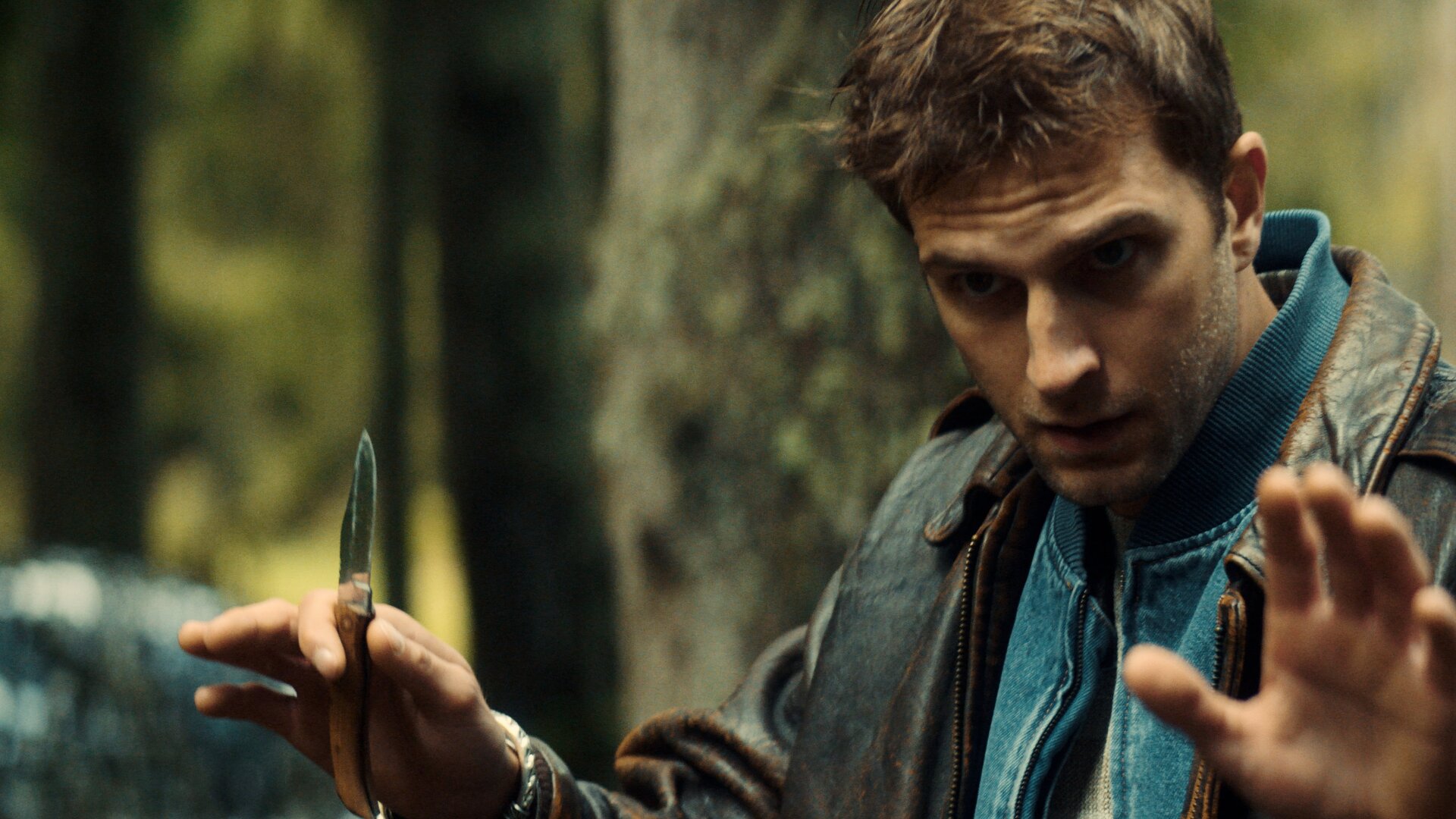 "Phoenix - Change Or Die: Die Macht der Aktionäre": Mathias Boissel (Léo Legrand) steht im Wald und hat seinen Blick nach vorne gerichtet, mit seinen Händen macht er eine beruhigende Geste, wobei er in seiner Rechten ein ausgeklapptes Taschenmesser hält.