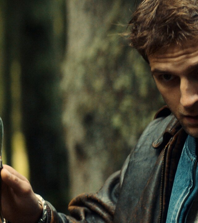 "Phoenix - Change Or Die: Die Macht der Aktionäre": Mathias Boissel (Léo Legrand) steht im Wald und hat seinen Blick nach vorne gerichtet, mit seinen Händen macht er eine beruhigende Geste, wobei er in seiner Rechten ein ausgeklapptes Taschenmesser hält.