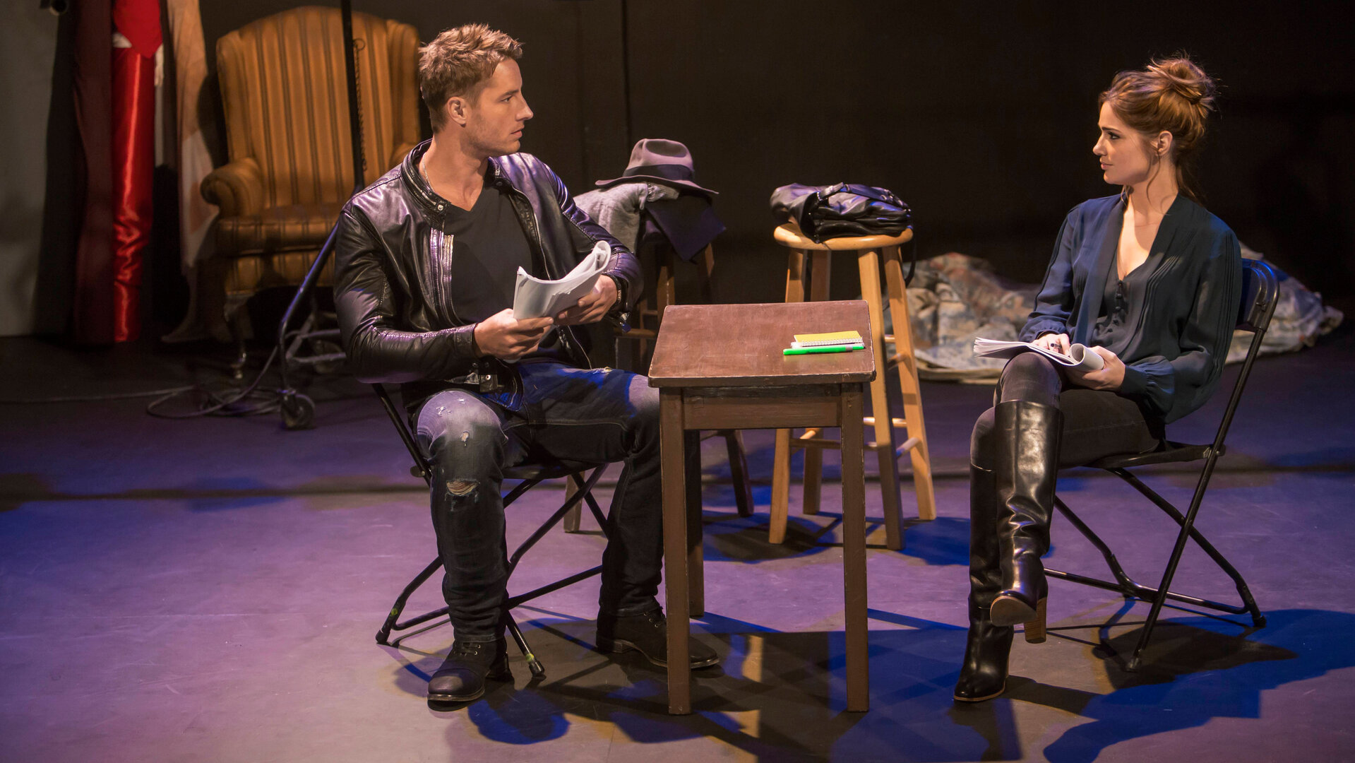 "This Is Us - Das ist Leben: Das magische T-Shirt": Kevin Pearson (Justin Hartley) und Olivia Maine (Janet Montgomery) sitzen sich auf einer Theaterbühne an einem schmalen Tisch gegenüber. Kevin hält ein Skript in den Händen und blickt Olivia fragend an. Olivia hält ebenfalls ein Skript in den Händen und blickt zu Kevin.