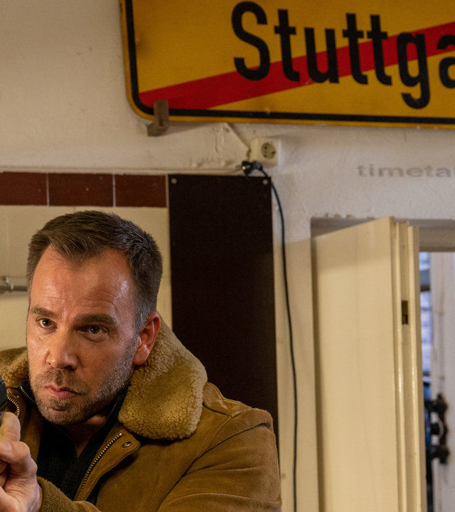"SOKO Stuttgart – Das Pfandhaus": Kriminaloberkommissar Lars Pöhlmann (Dominc Boeer) steht mit gezogener Dienstwaffe in der Küche einer Stuttgarter Wohnung. Mit festem Blick fokussiert er sein Gegenüber, welches nicht zu sehen ist. Hinter ihm hängt an der Wand ein Stuttgarter Ortsausgangsschild.