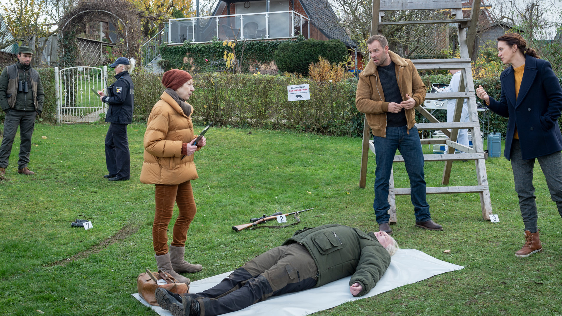 "SOKO Wismar – Waidmannsheil": Eine Wiese in einer Kleingartenkolonie. Die Leiche von Hubertus Voss (Andreas Euler) liegt auf einer Plane. Rechts davon stehen Lars Pöhlmann (Dominic Boeer) und Karoline Joost (Nike Fuhrmann), die zu Dr. Helene Sturbeck (Katharina Blaschke) sehen. Weiter links spricht Jens Kemper (Jörg Kunze) mit einer Polizistin. Jedes Beweismittel ist mit je einem Schild gekennzeichnet: 7.) ein Fernglas, 2.) ein Scharfschützengewehr und 3.) ein mobiler Ansitz.