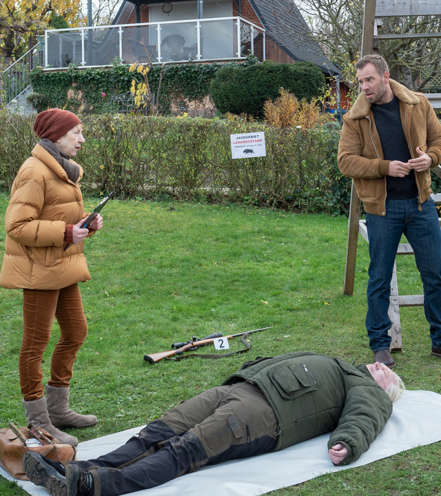 "SOKO Wismar – Waidmannsheil": Eine Wiese in einer Kleingartenkolonie. Die Leiche von Hubertus Voss (Andreas Euler) liegt auf einer Plane. Rechts davon stehen Lars Pöhlmann (Dominic Boeer) und Karoline Joost (Nike Fuhrmann), die zu Dr. Helene Sturbeck (Katharina Blaschke) sehen. Weiter links spricht Jens Kemper (Jörg Kunze) mit einer Polizistin. Jedes Beweismittel ist mit je einem Schild gekennzeichnet: 7.) ein Fernglas, 2.) ein Scharfschützengewehr und 3.) ein mobiler Ansitz.