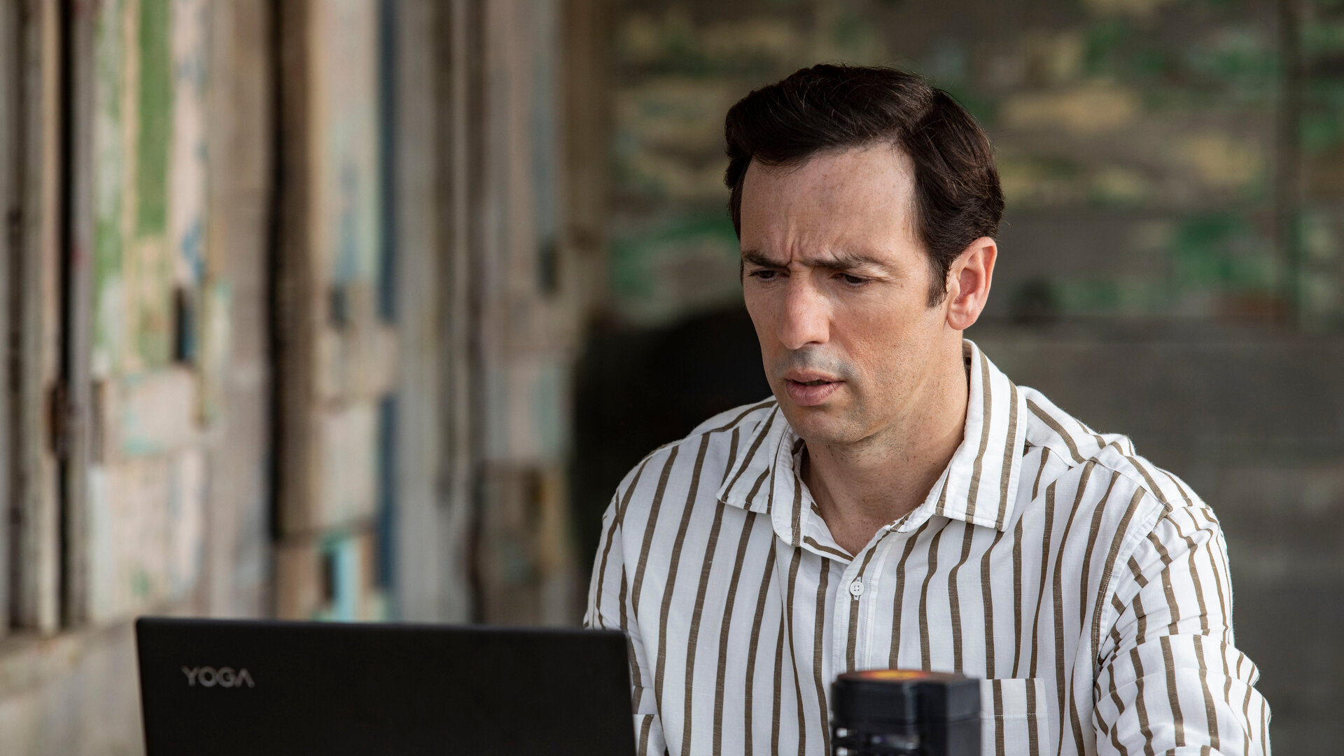 "Death in Paradise - Wechselstrom": DI Neville Parker (Ralf Little) sitzt mit seinem Laptop an einem Tisch und blickt konzentriert und leicht besorgt auf den Bildschirm.