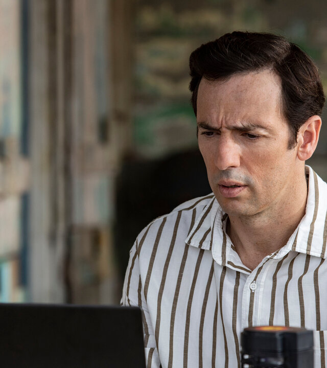 "Death in Paradise - Wechselstrom": DI Neville Parker (Ralf Little) sitzt mit seinem Laptop an einem Tisch und blickt konzentriert und leicht besorgt auf den Bildschirm.