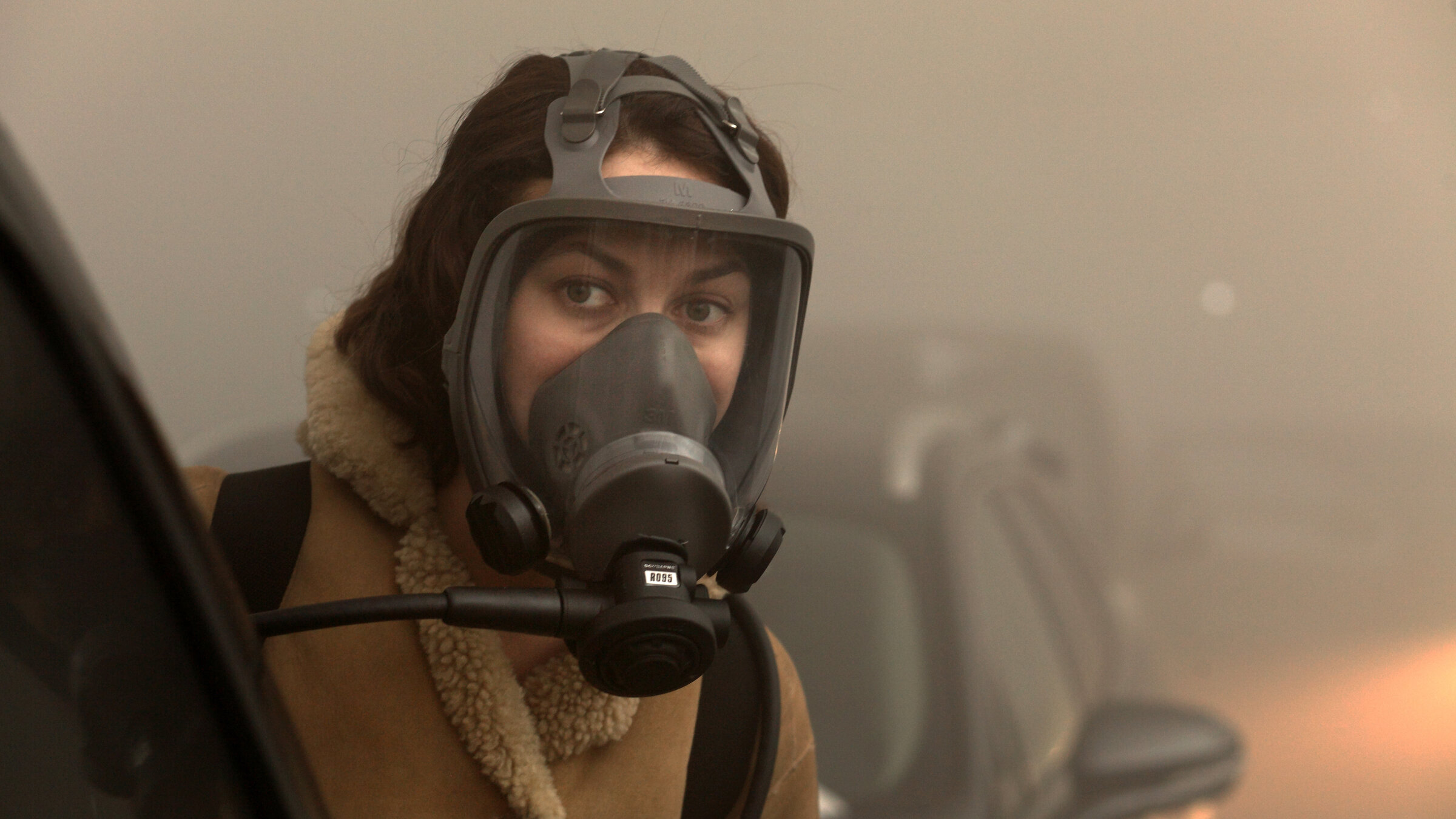 "A Breath Away": Nahaufnahme: Anna (Olga Kurylenko) steht mit einer Atemschutzmaske in einer vernebelten Straße zwischen zwei Autos.