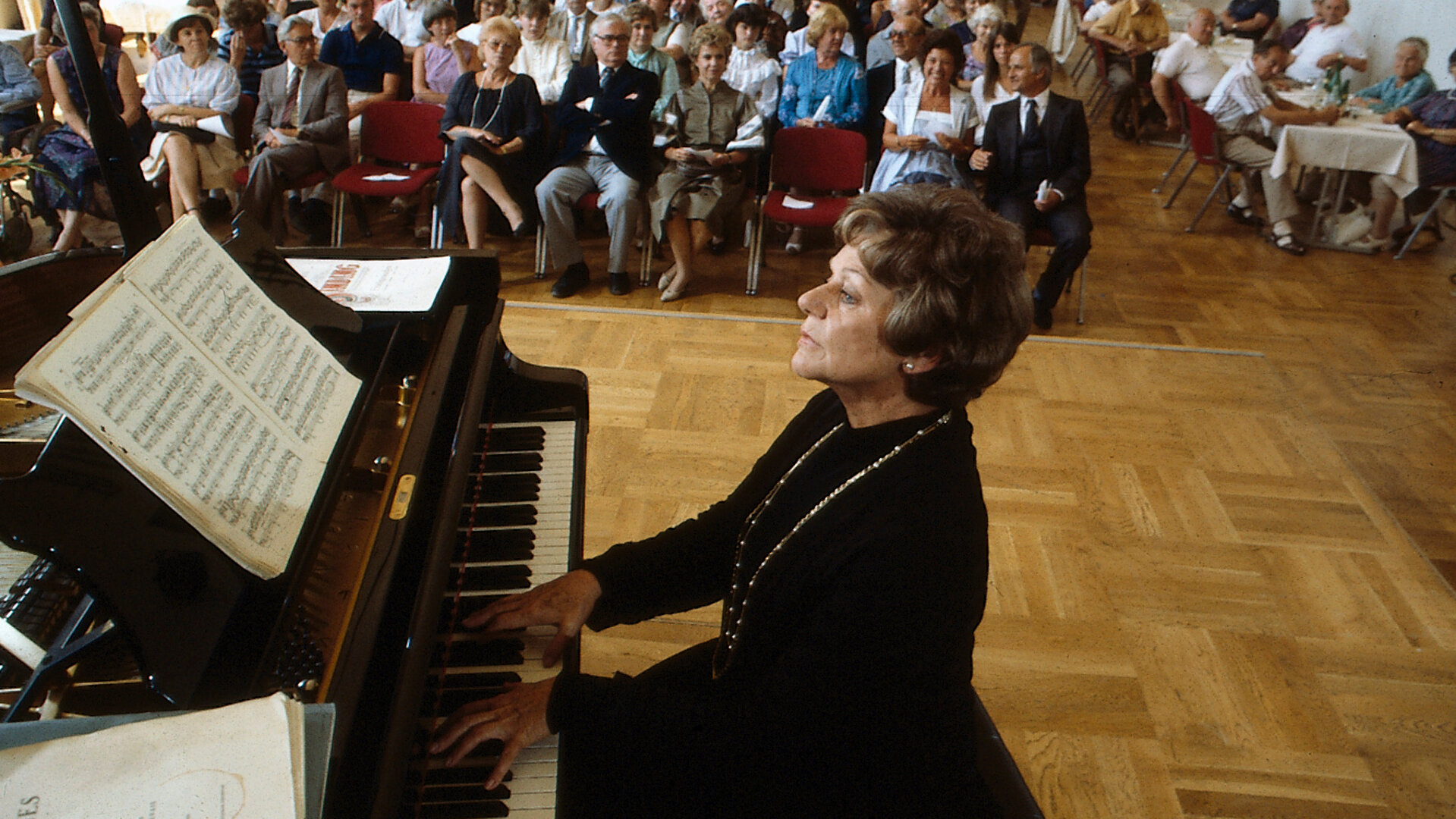 "Diese Drombuschs -  Das Konzert": Oma Drombusch (Grete Wurm) gibt ein Klavierkonzert vor einem größeren Publikum in einem Saal.