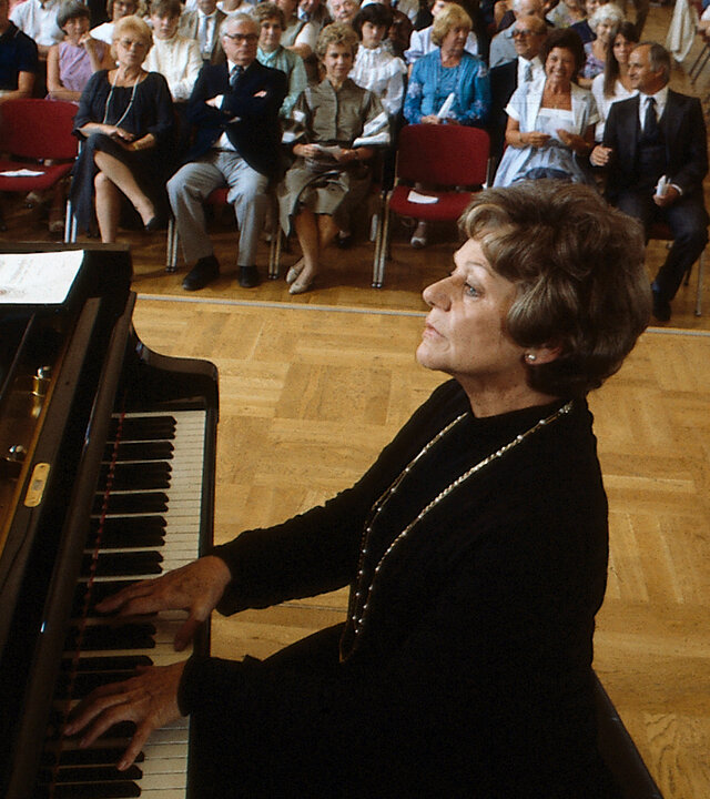 "Diese Drombuschs -  Das Konzert": Oma Drombusch (Grete Wurm) gibt ein Klavierkonzert vor einem größeren Publikum in einem Saal.