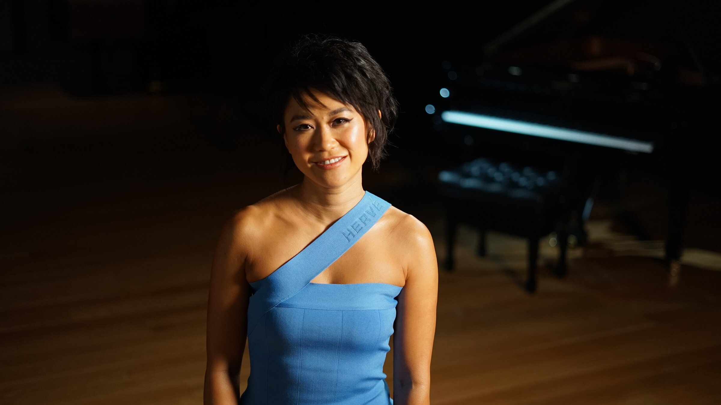 "Konzert der Wiener Philharmoniker aus Paris": die chinesische Pianistin Yuja Wang steht auf einer Bühne und lächelt in die Kamera. Sie trägt ein trägerloses hellblaues Abendkleid. Im Hintergrund ein schwarzer Flügel.