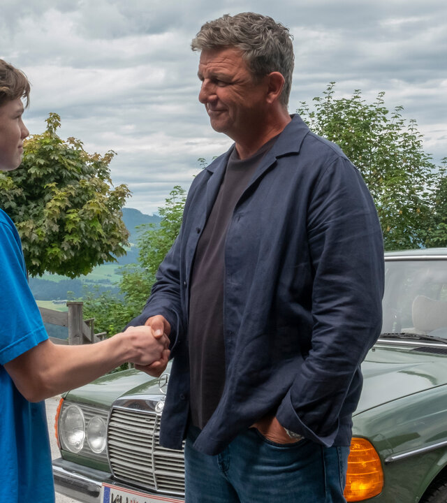 "Der Bergdoktor: Kindeswohl": Vor dem Haus der Familie Fischer: Vor dem grünen Mercedes stehen sich Dr. Martin Gruber (Hans Sigl) und John Luckau (Nico Marischka) gegenüber und reichen sich die Hände. Dr. Gruber lächelt den Jungen aufmunternd an.