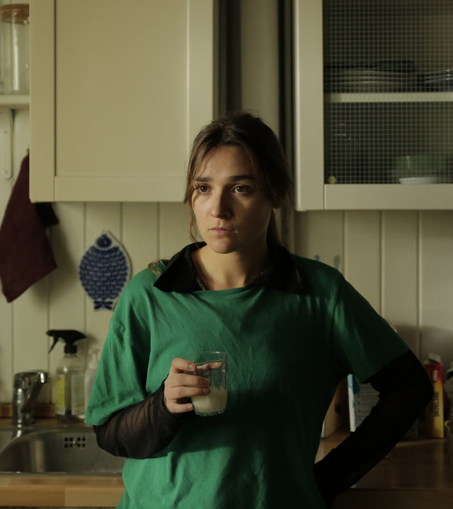 "About Saturday - Forever Singles": Klara (Darin Hagi) steht in einer Küche und schaut konzentriert in die Ferne, während sie ein Glas Milch in der Hand hält.