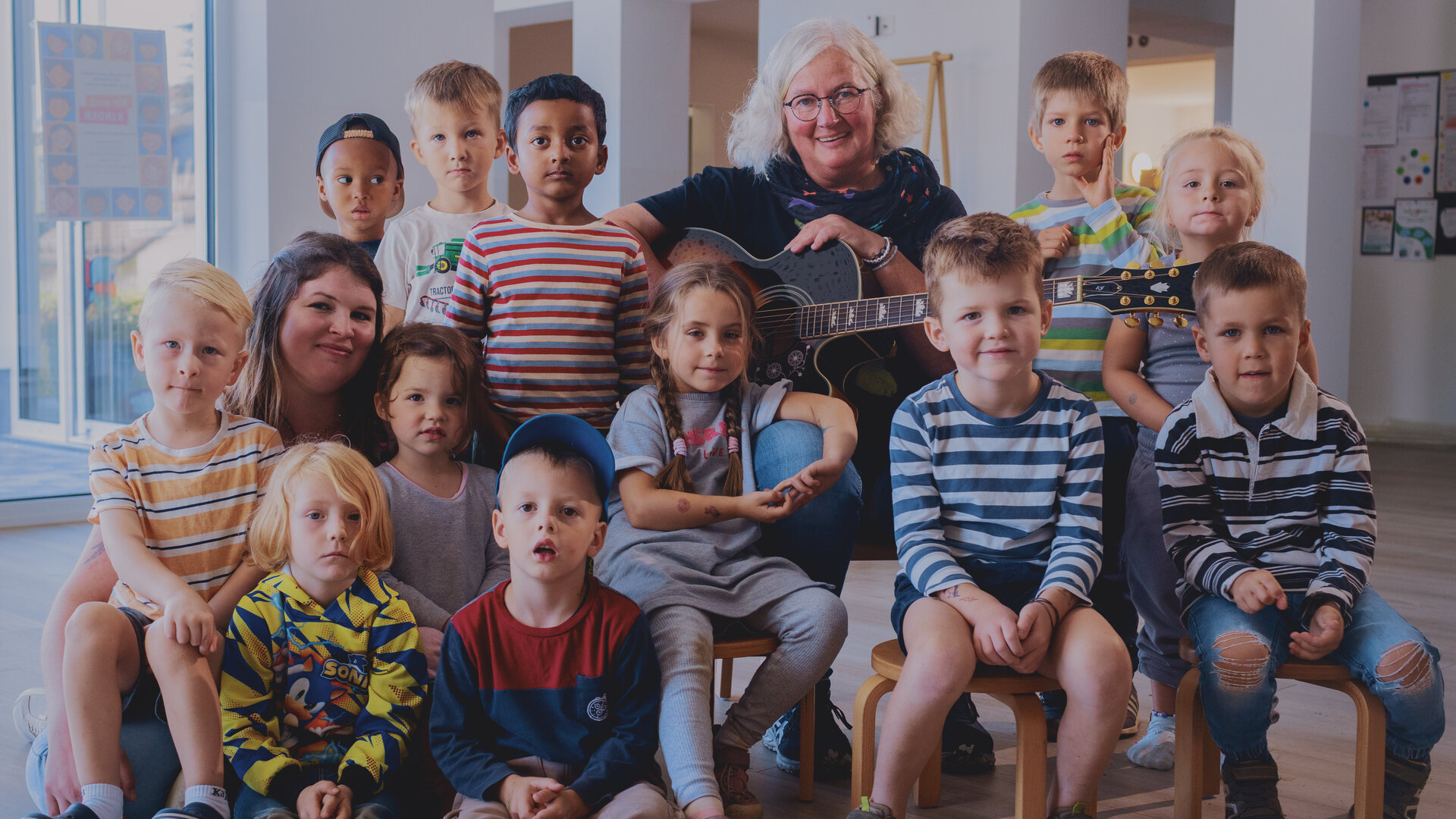 "Aktion Mensch - Glückszahlen der Woche": Eine Gruppe von Kindern sitzt in einem hellen Raum auf kleinen Stühlen, eine erwachsene Person sitzt bei ihnen und hält eine Gitarre. Alle schauen freundlich in die Kamera.