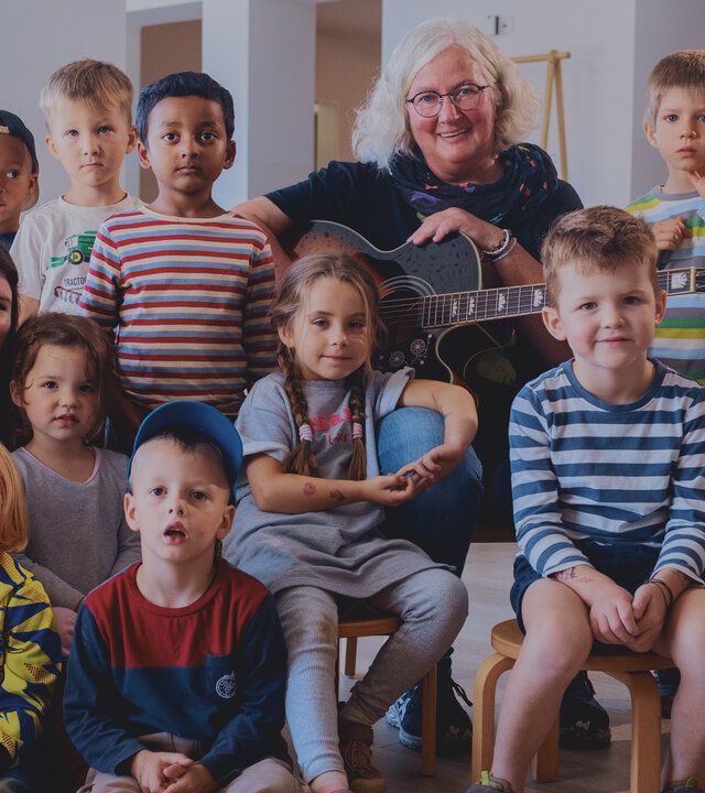 "Aktion Mensch - Glückszahlen der Woche": Eine Gruppe von Kindern sitzt in einem hellen Raum auf kleinen Stühlen, eine erwachsene Person sitzt bei ihnen und hält eine Gitarre. Alle schauen freundlich in die Kamera.