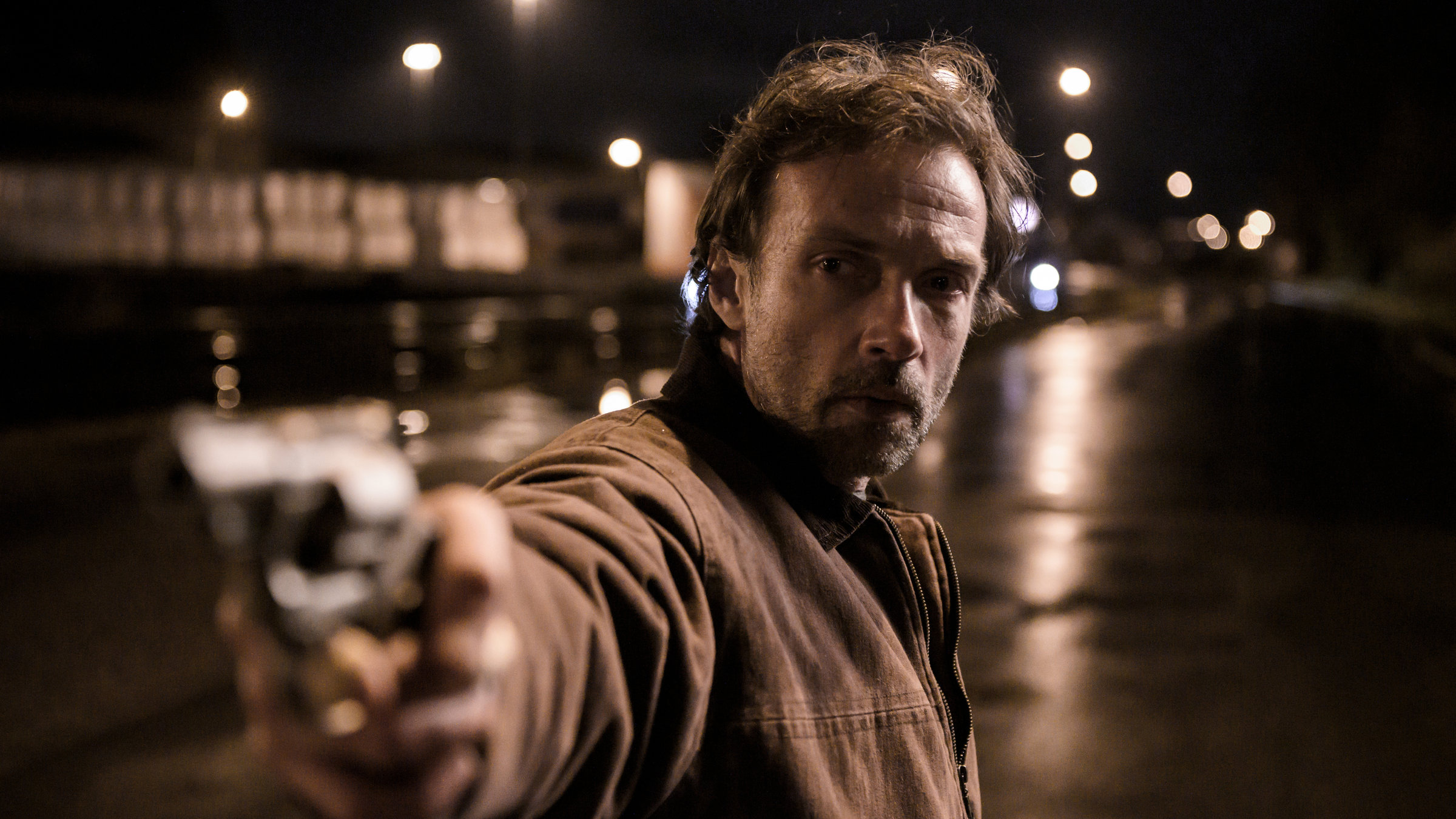 "Die Zukunft ist ein einsamer Ort": Frank (Lucas Gregorowicz) zielt mit einem Revolver und blickt gleichzeitig auf ein Ziel neben der Kamera. Er steht nachts auf einer regennassen Straße.