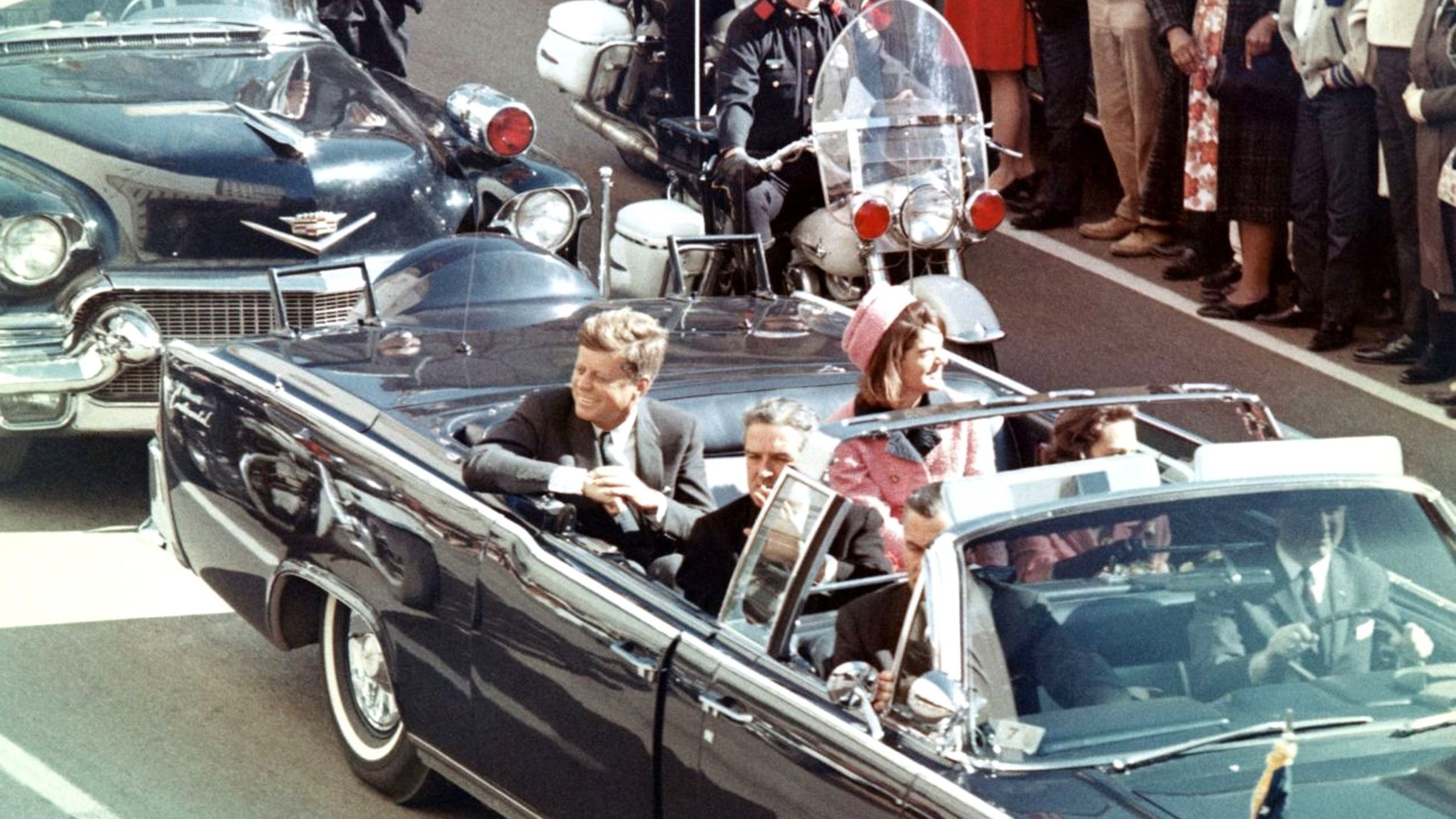 "JFK Revisited - Die Wahrheit über den Mord an John F. Kennedy": Zwei Männer und eine Frau sitzen lachend und winkend in einem dachlosen Auto während die Menge ihnen zujubelt.