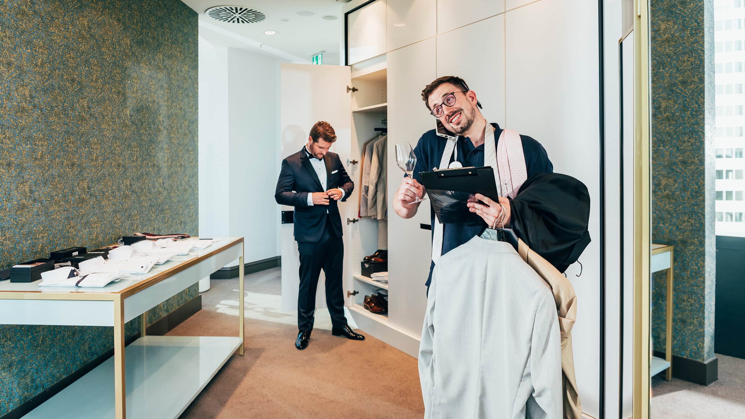 "37°Leben - Stresstest Trauzeug*in: Zwischen Party, Planung und Prosecco": Lukas steht mit Klemmbrett und Sektglas in der Hand sowie Anzügen auf dem Arm in einem Wohnraum. Er hat ein Handy zwischen Ohr und Schultern geklemmt. Hinter ihm steht Maurice vor einer offenen Schranktür und knöpft sein Jackett zu.