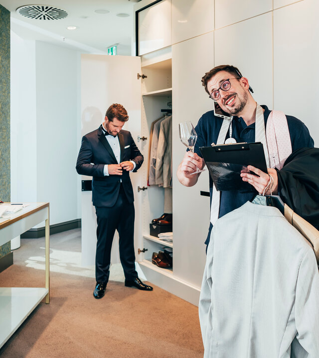 "37°Leben - Stresstest Trauzeug*in: Zwischen Party, Planung und Prosecco": Lukas steht mit Klemmbrett und Sektglas in der Hand sowie Anzügen auf dem Arm in einem Wohnraum. Er hat ein Handy zwischen Ohr und Schultern geklemmt. Hinter ihm steht Maurice vor einer offenen Schranktür und knöpft sein Jackett zu.