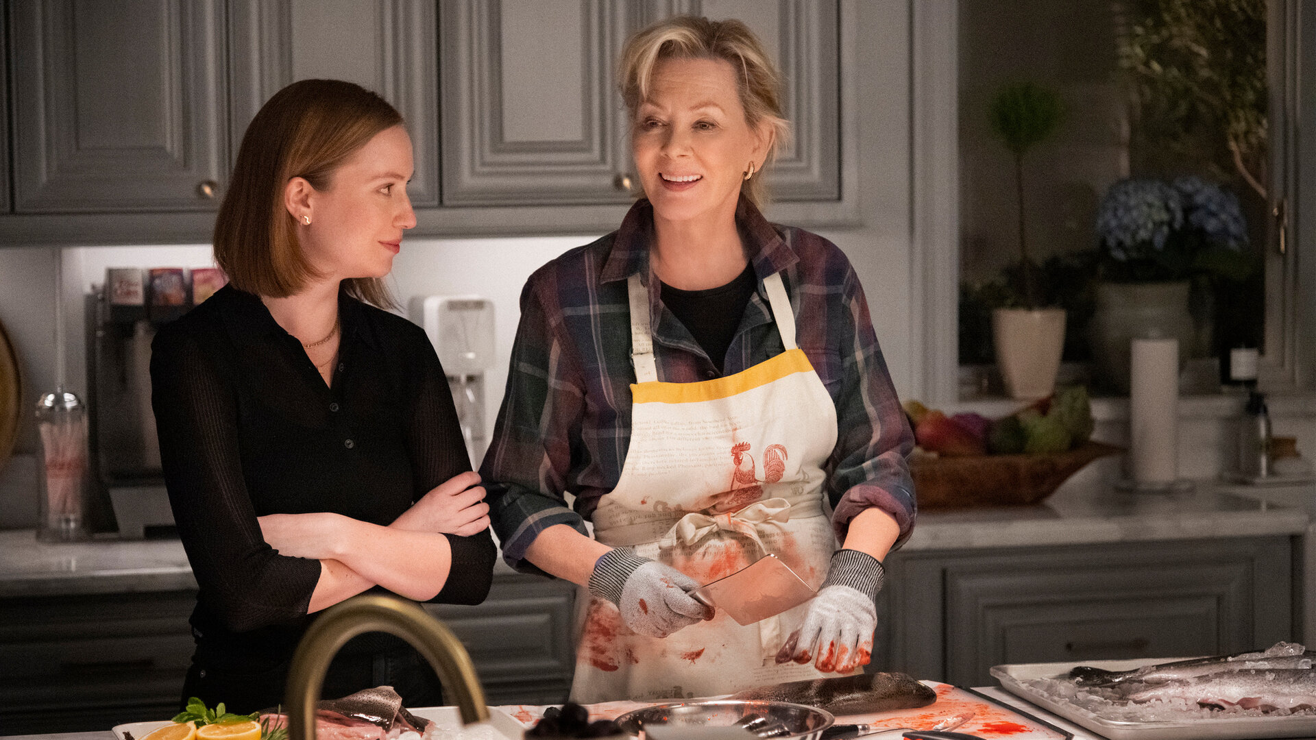 "Hacks - Show, die letzte": Ava (Hannah Einbinder) steht mit verschränkten Armen neben Deborah (Jean Smart) in einer Küche und blickt zu ihr, während Deborah eine blutbefleckte Schürze trägt, mit Handschuhen ein Hackmesser hält und dabei lächelnd in ihre Richtung schaut.