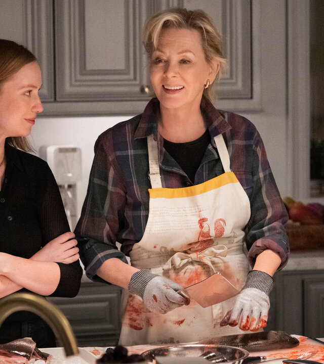 "Hacks - Show, die letzte": Ava (Hannah Einbinder) steht mit verschränkten Armen neben Deborah (Jean Smart) in einer Küche und blickt zu ihr, während Deborah eine blutbefleckte Schürze trägt, mit Handschuhen ein Hackmesser hält und dabei lächelnd in ihre Richtung schaut.