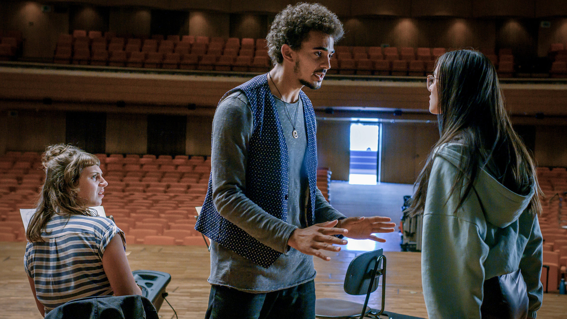 "SOKO Linz - Da Capo": Bernardo Souza (Leonard Burkhardt) und Nancy Liu (Julia Strowski) diskutieren während der Probe auf der Bühne. Adina Morosan (Josephine Bloéb) beobachtet entsetzt das Geschehen. Im Hintergrund sind die unteren und oberen Ränge des Saals zu sehen.