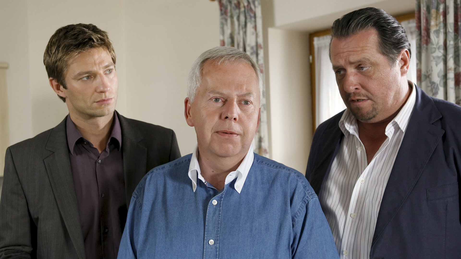 "Die Rosenheim-Cops - Urlaubsreise in den Tod": Kommissar Hansen (Igor Jeftic), Karl Hinrichs (Harald Maack) und Kommissar Prantl (Andreas Giebel) stehen in einem Wohnraum. Herr Hinrichs schaut nach vorne, die Kommissare bobachten ihn.