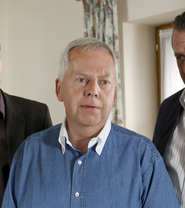 "Die Rosenheim-Cops - Urlaubsreise in den Tod": Kommissar Hansen (Igor Jeftic), Karl Hinrichs (Harald Maack) und Kommissar Prantl (Andreas Giebel) stehen in einem Wohnraum. Herr Hinrichs schaut nach vorne, die Kommissare bobachten ihn.
