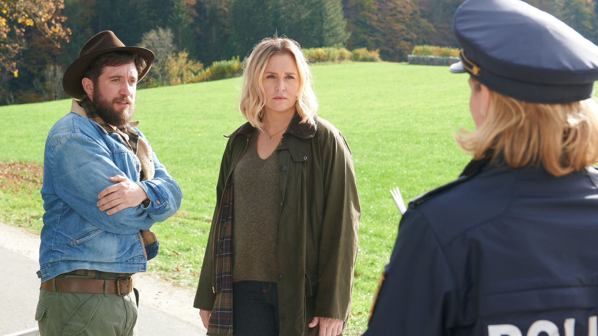 "Lena Lorenz - Gegen den Schmerz": Auf einer Straße stehen Schorschi Striebel (Sebastian Edtbauer) und Lena Lorenz (Judith Hoersch) und reden mit der Polizeibeamtin Anna Prückl (Sarah-Lavinia Schmidbauer), die von hinten zu sehen ist. Schorschi hat die Arme um seinen Oberkörper geschlungen und wirkt schockiert. Lena schaut die Polizistin ernst an.