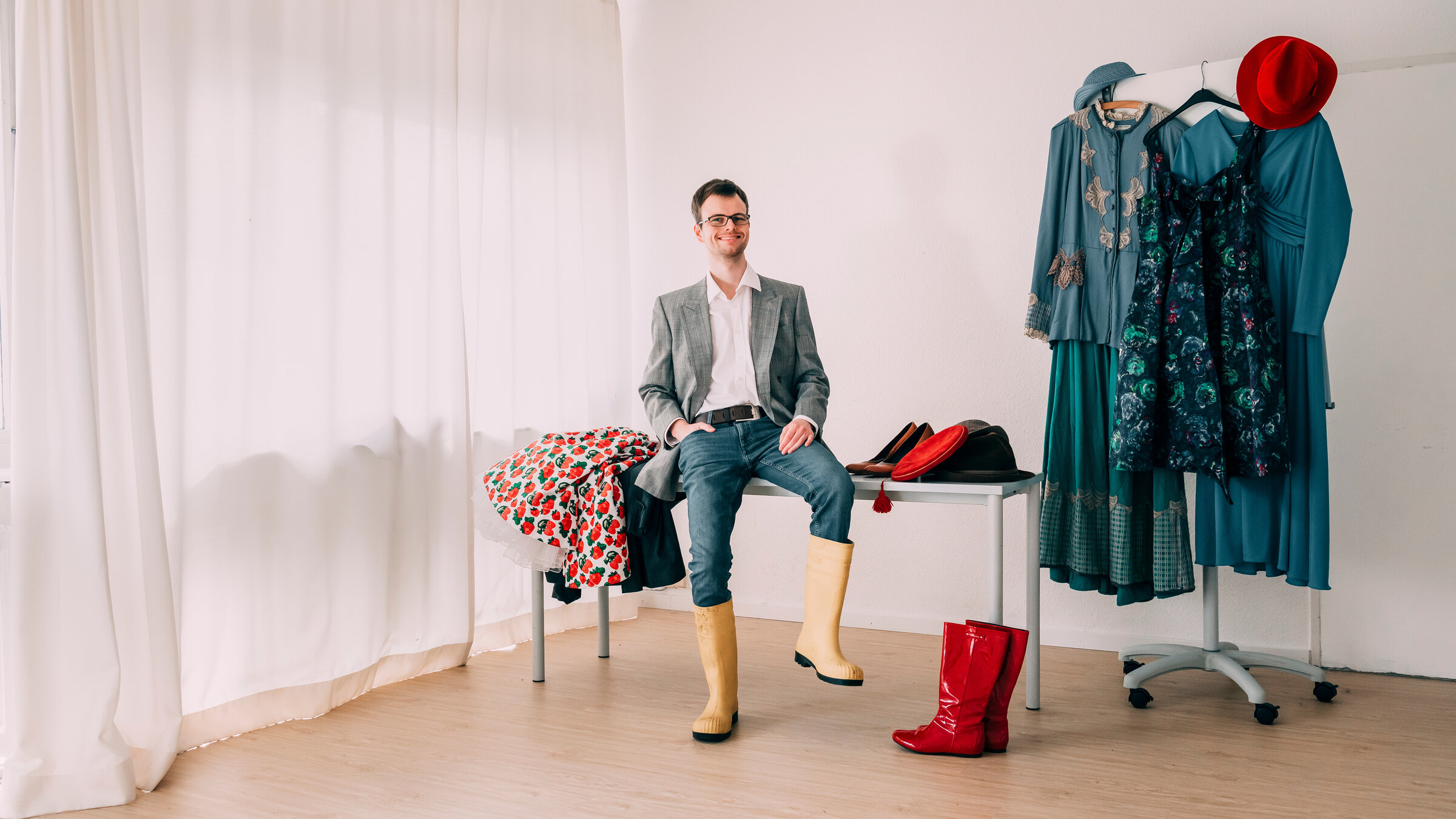 "Einfach Mensch - Werkstatt, ohne mich": Rasmus sitzt in einem hellen Raum auf einem Tisch auf dem Kleidung liegt. Darunter steht ein Paar rote Stiefel. Daneben steht auf der einen Seite ein Kleiderständer mit blauen Kleidern und einem roten Hut; auf der anderen Seite ist ein Fenster, das mit einem weißen Vorhang verdeckt ist. Rasmus lächelt in die Kamera.