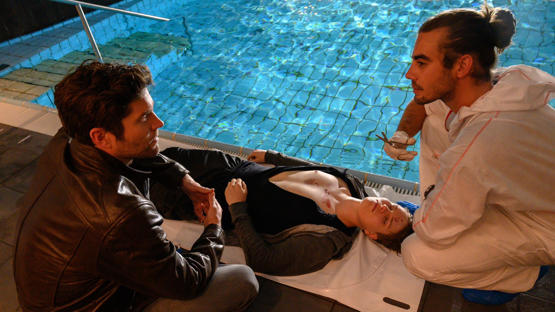 "SOKO Stuttgart - Kranke Liebe": Jo Stoll (Peter Ketnath) und Benedikt Förster (Florian Wünsche) sitzen neben dem Pool in einer Rehaklinik in der Hocke. Zwischen ihnen liegt die in einen Bademantel gehüllte Leiche von Paul Steinfeld (Stefan Siewert).
